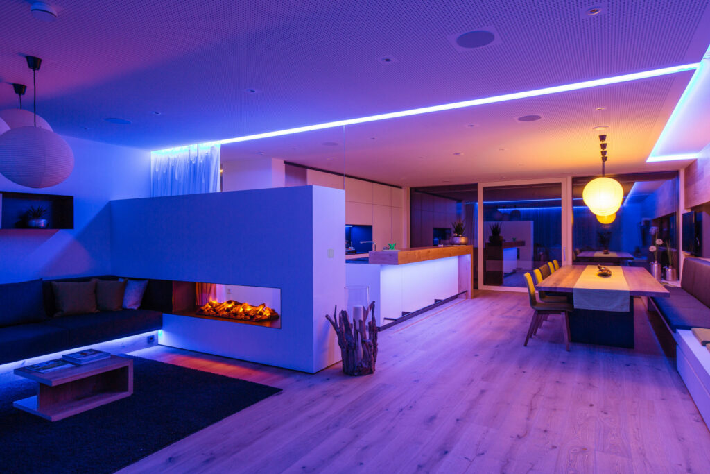 Wohnraum am Abend mit indirekter Beleuchtung in Blau und Violett, Kamin, Küche und Esstisch